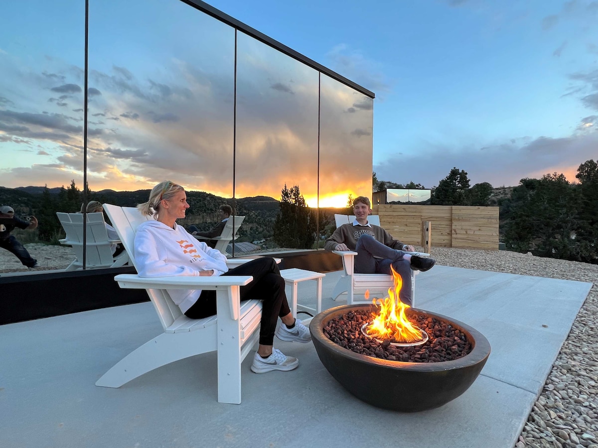 Two individuals enjoy a peaceful evening by a fire pit, seated in white Adirondack chairs. The high mountains and colorful sunset are reflected in the mirrored exterior of the house, complementing the serene setting.