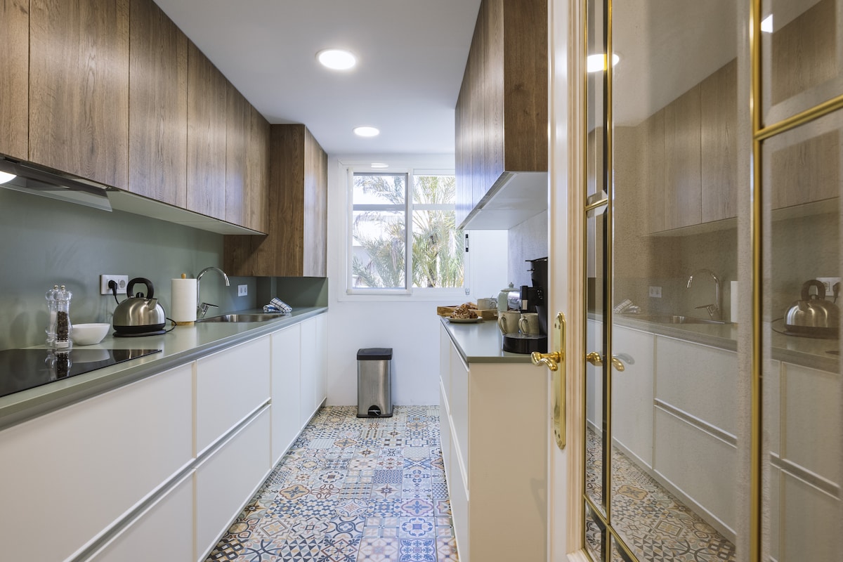 A modern kitchen is showcased with sleek cabinetry and an open layout. Essential appliances are positioned for convenience, while a window allows natural light to illuminate the space. Colorful patterned tiles add visual interest to the floor, enhancing the room's functionality.