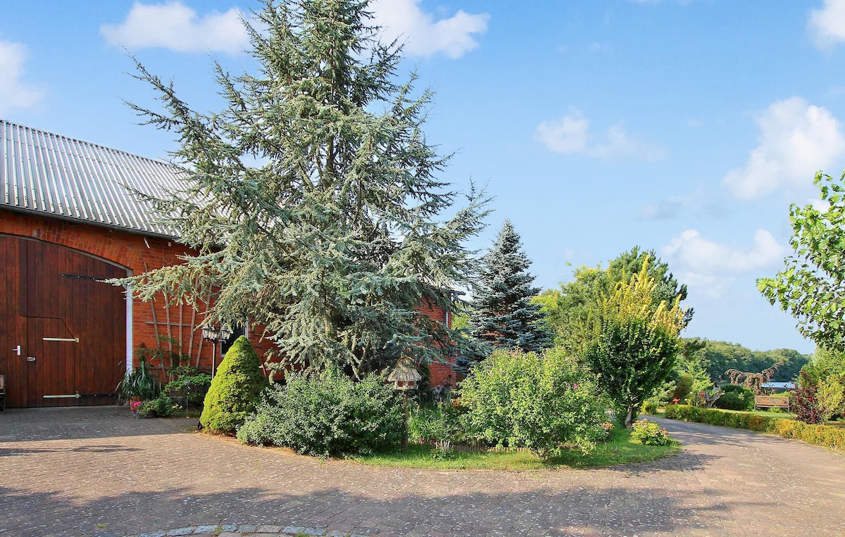 A well-maintained garden area is showcased, featuring a mix of shrubs and trees, including a large conifer. A paved pathway leads to the entrance of a former barn, surrounded by greenery and flowers under a clear blue sky.