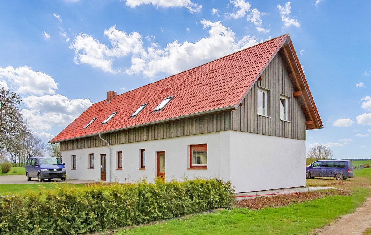The exterior of the renovated barn features a combination of wooden paneling and white walls, topped with a distinctive red roof. Windows are placed symmetrically, allowing natural light into the building. A neatly trimmed hedge lines the front, with a gravel pathway leading to the entrance.