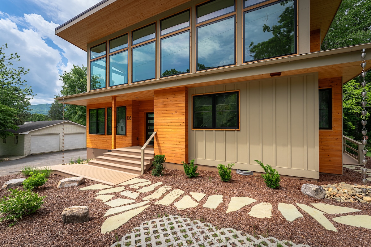 The entrance to a modern home is showcased, featuring a wooden facade and large glass windows that allow natural light to fill the interior. Stone pathways lead to the front steps, which are complemented by landscaped greenery and a gravel area, enhancing the inviting exterior.