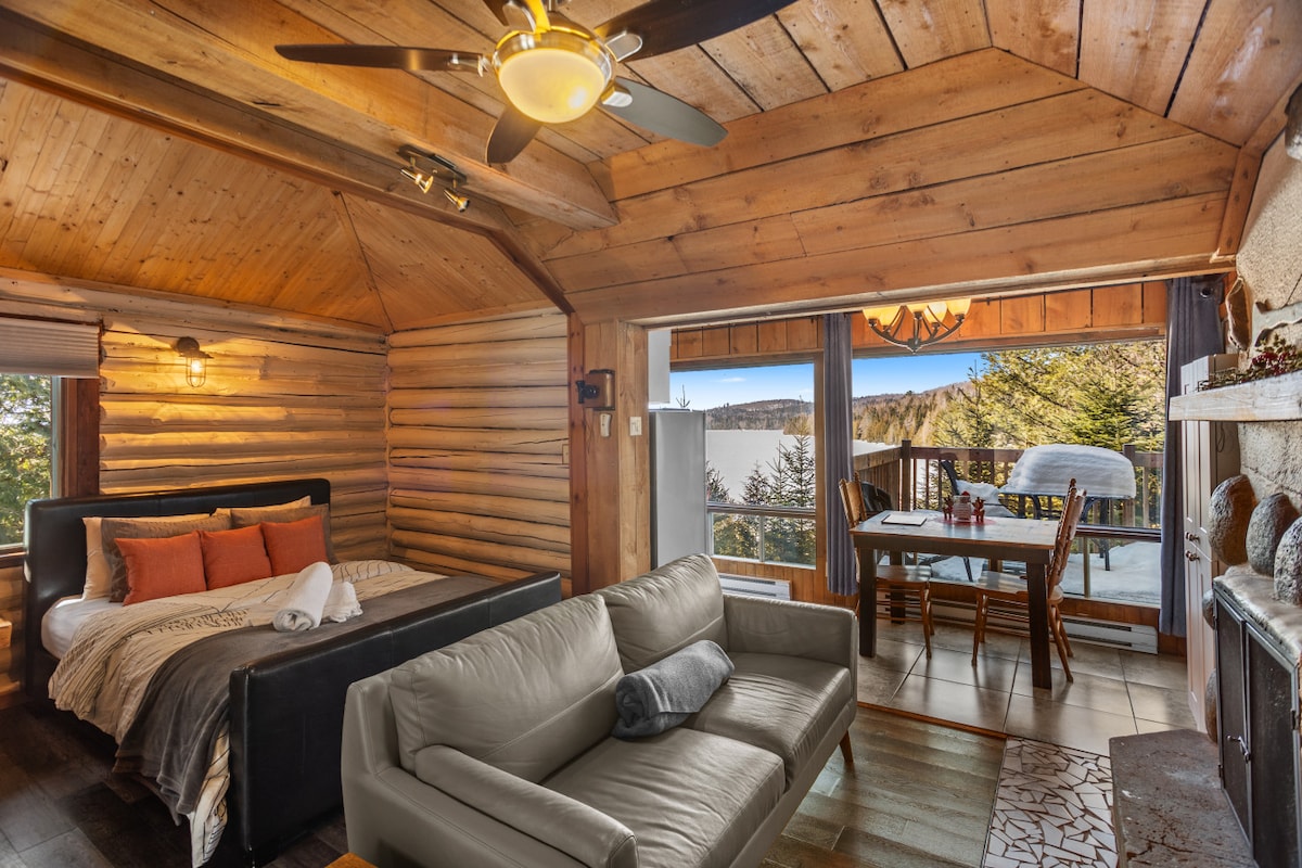 A cozy loft-style indoor space features a spacious bed with soft linens, complemented by a stylish sofa and a dining area overlooking the scenic landscape through large glass doors. Warm wood paneling adds to the rustic charm, while natural light floods the room.