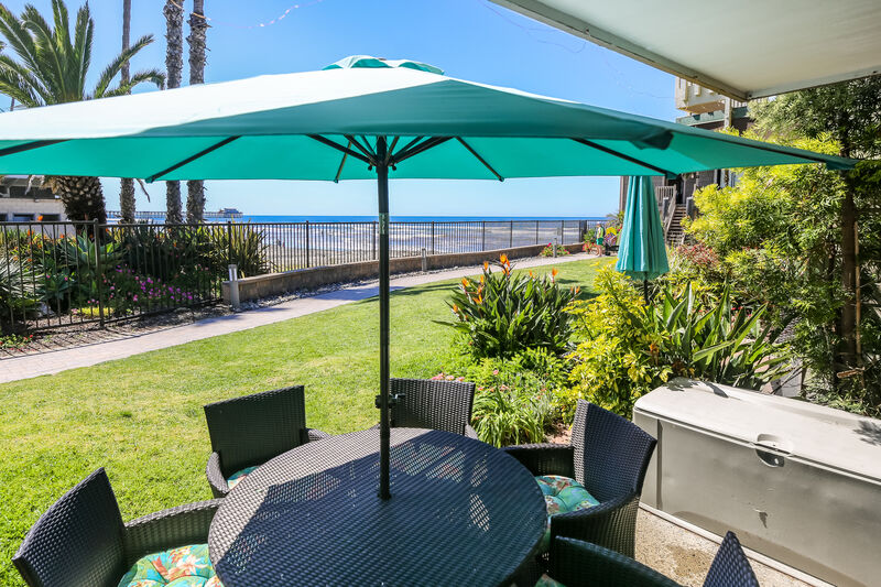 A patio area is visible with a round table surrounded by six dark wicker chairs, brightly cushioned for comfort. Above, a large turquoise umbrella offers shade, while lush green grass and colorful landscaping extend towards the ocean view beyond a gated perimeter.