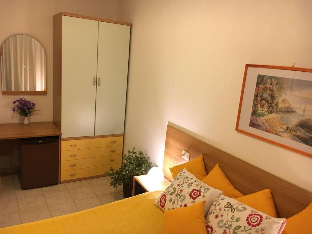 A cozy bedroom is highlighted by a spacious bed adorned with colorful pillows. A wooden wardrobe and a desk are situated nearby, while a mirror reflects light from the wall. A small plant adds a touch of greenery, and artwork decorates the wall.