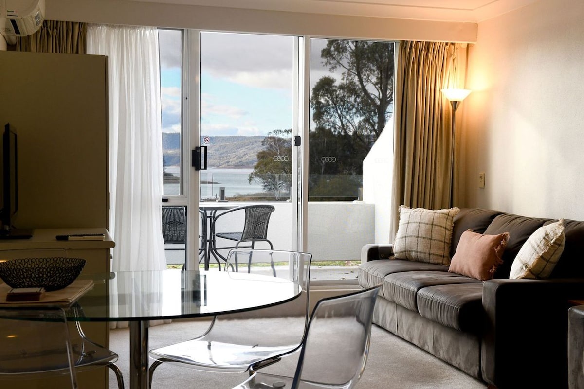 A living and dining area is visible, featuring a comfortable sofa and a glass dining table. Large sliding doors provide views of the outdoor space, with trees and water in the background. Soft lighting from a floor lamp complements the neutral decor.