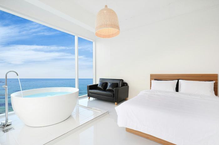 A bright and airy room features a spacious bed alongside a black leather sofa. A large window offers an unobstructed view of the sea. A stylish freestanding bathtub sits near the window, enhancing the tranquil atmosphere of the space.