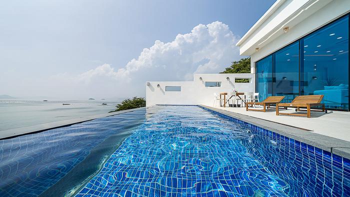 A modern infinity pool stretches along the terrace, offering clear views of the sea. Sun loungers are positioned near the water, reflecting bright blue tiles that harmonize with the sky. Large glass windows invite natural light into the space, enhancing the serene atmosphere.