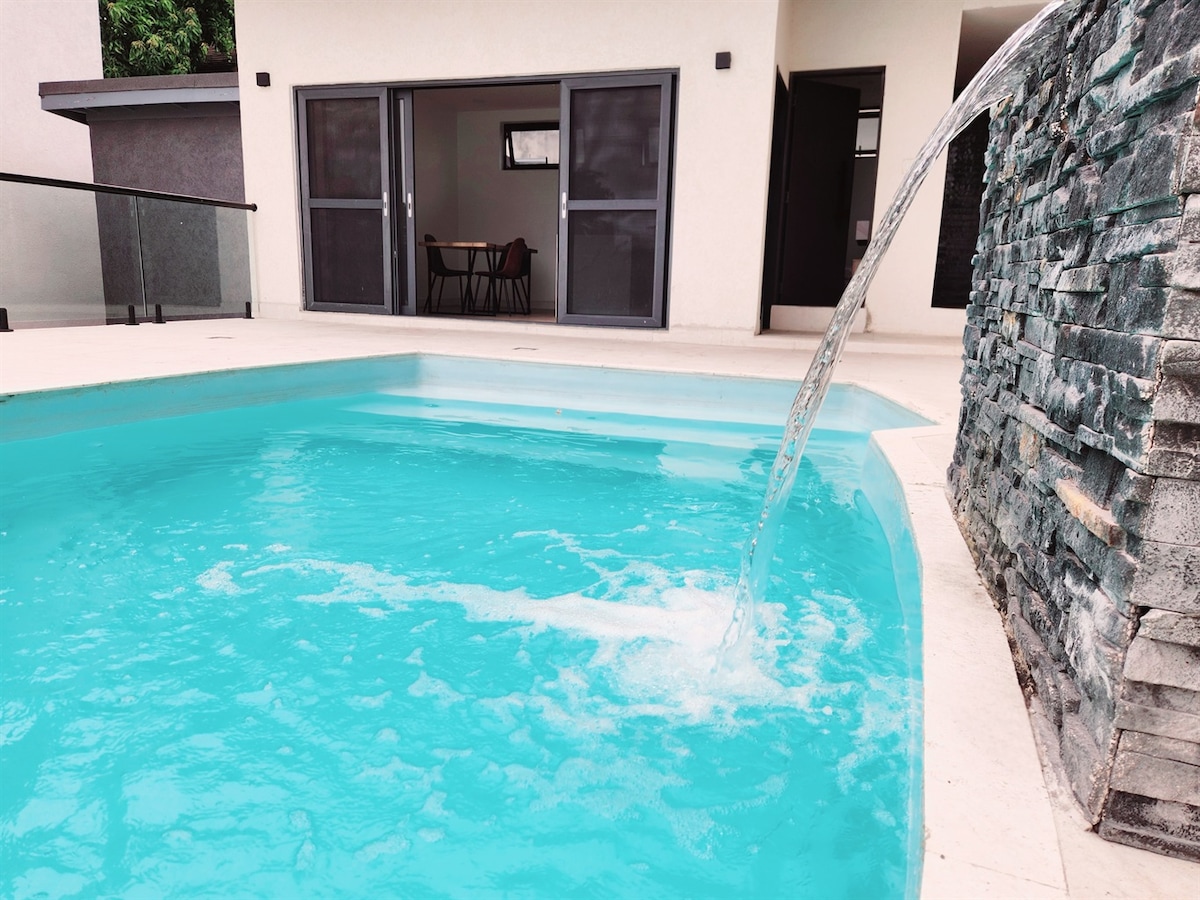 A refreshing pool is visible, featuring a water feature that flows into the turquoise water. The pool area is surrounded by sleek stonework and leads to a modern entrance with glass doors, allowing for a seamless connection with the living space.