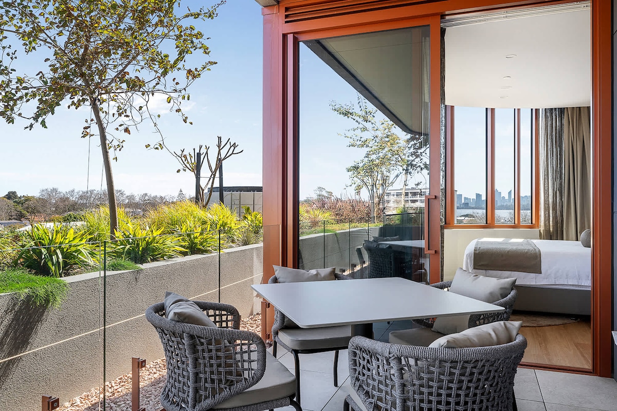 A private balcony is adorned with modern wicker chairs surrounding a light table. Large glass doors allow natural light to flow into the living space, revealing a tranquil view of greenery and the distant city skyline.
