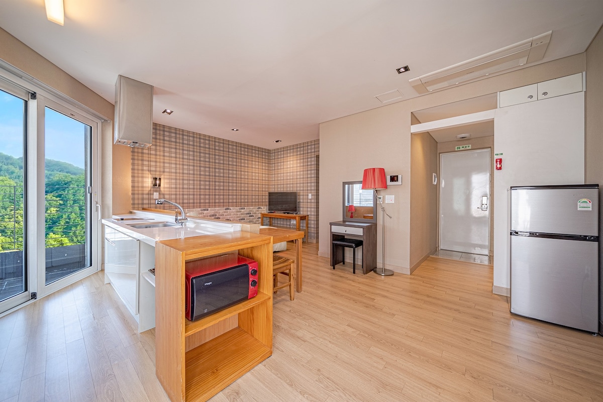 An open living area is presented, featuring a modern kitchen with an island and wooden dining table. Large windows provide views of the lush landscape, while light wood flooring complements the neutral wall tones. A cozy seating area includes a small desk and a stylish lamp.