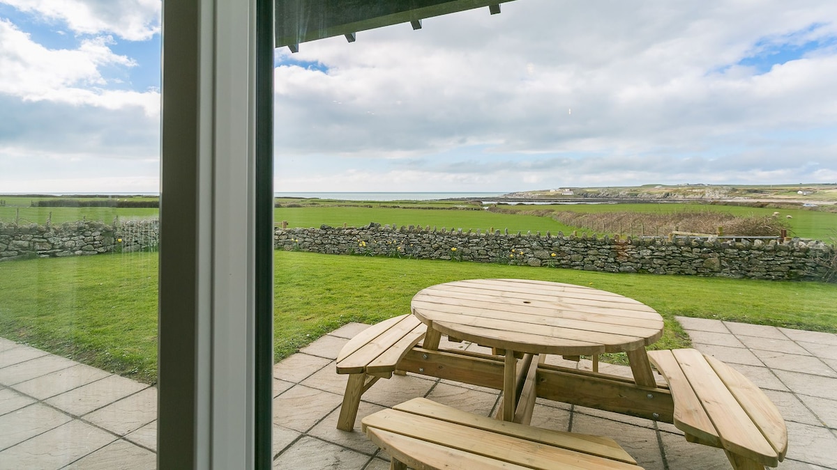 A round picnic table made of wood is set on a paved patio, offering expansive views of green fields and the sea in the distance. Stone walls outline the property, with a cloudy sky above casting natural light across the scene.
