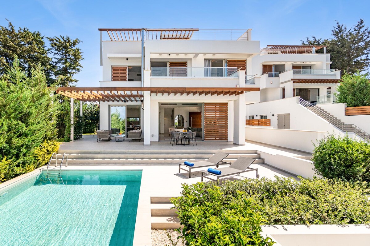 A modern villa exterior showcases a spacious terrace with comfortable lounge chairs and a private pool. Lush greenery surrounds the area, providing a tranquil setting. The building features multiple levels and large windows, inviting ample natural light and offering views of the well-maintained landscape.