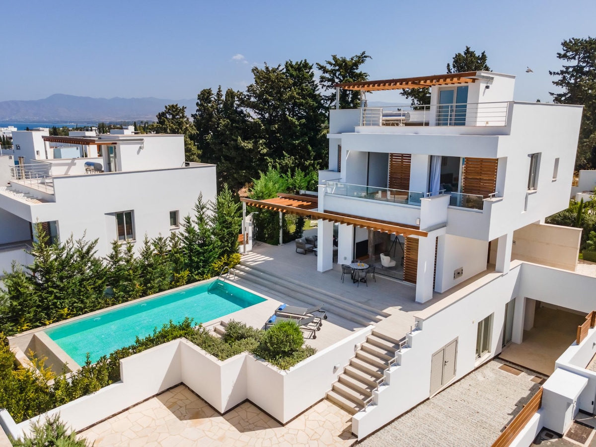 An aerial view of a modern villa showcases a private swimming pool, surrounded by landscaped greenery. The spacious outdoor area features a covered terrace with outdoor seating, while the villa’s sleek white architecture contrasts with the vibrant natural surroundings.
