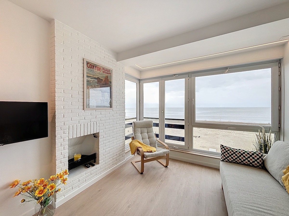 A comfortable living area features large windows showcasing expansive sea views. A light-colored sofa is positioned near the window, accompanied by a wooden chair. A minimalistic fireplace framed by a white brick wall adds to the space's welcoming ambiance.