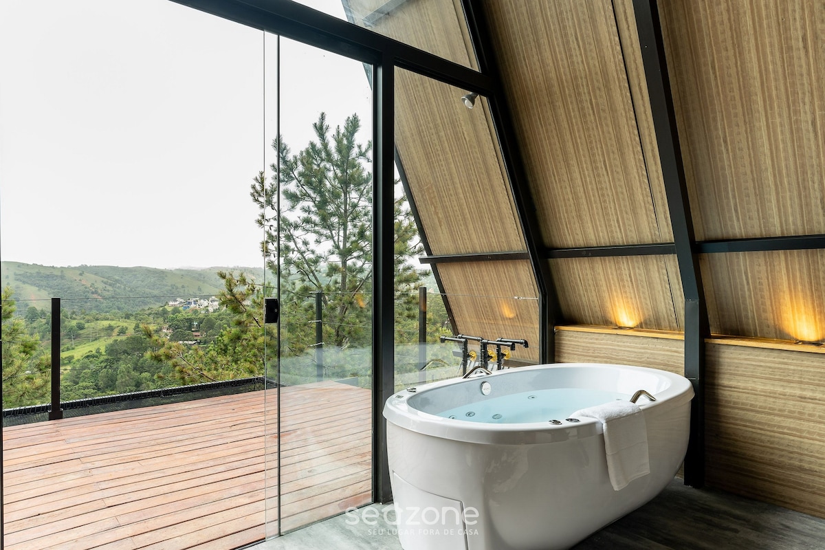 A modern private jacuzzi is positioned beside large glass doors, offering sweeping views of the surrounding mountains and forests. Natural wood accents complement the sleek design, while soft lighting creates a calming ambiance. A spacious wooden deck extends outside for additional relaxation.