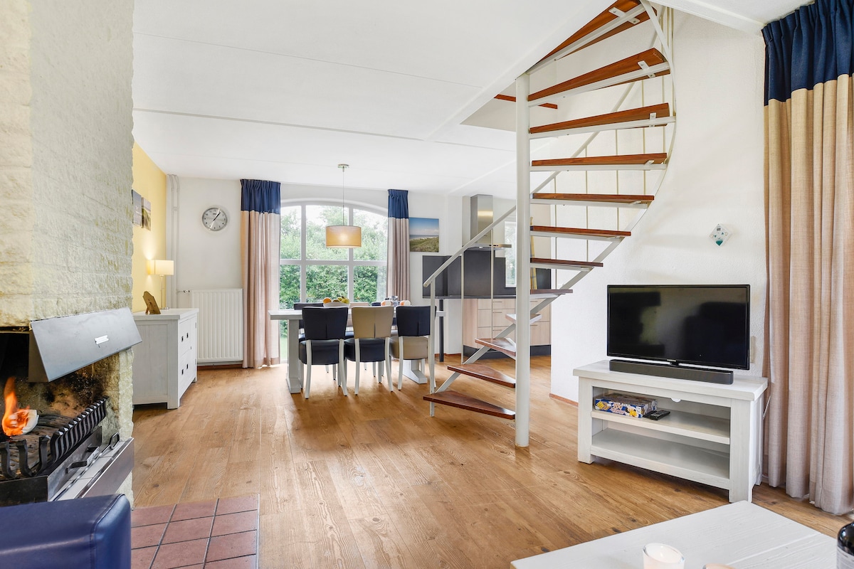 An open living space is presented with a spiral staircase leading to the upper floor. A seating area with a television is positioned beside a fireplace, while a dining table accommodates four chairs in the background. Natural light filters through large windows, creating a welcoming environment.
