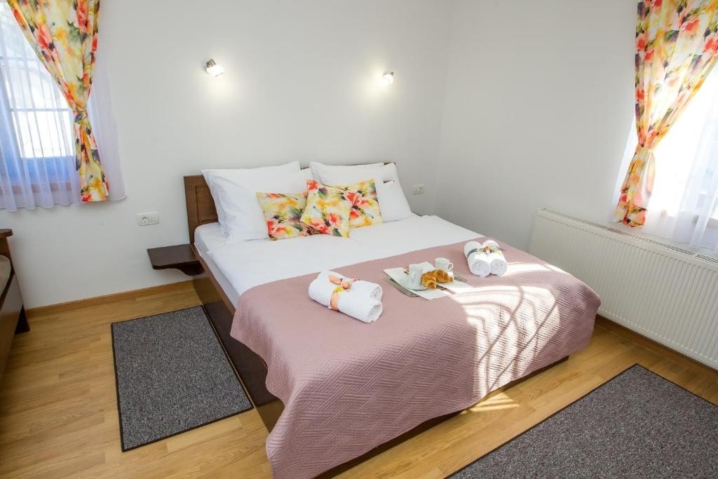 A sleeping area features a spacious bed with a soft pink quilt. Colorful floral curtains frame the windows, allowing natural light to brighten the room. A small bedside table holds a breakfast tray with pastries and two rolled towels are neatly placed at the foot of the bed.