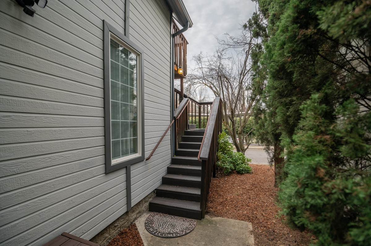 Sturdy wooden steps lead up to the second floor entrance of the loft, flanked by greenery. A large window with divided panes allows light to enter, while a small mat is situated on the landing, creating a welcoming first impression.