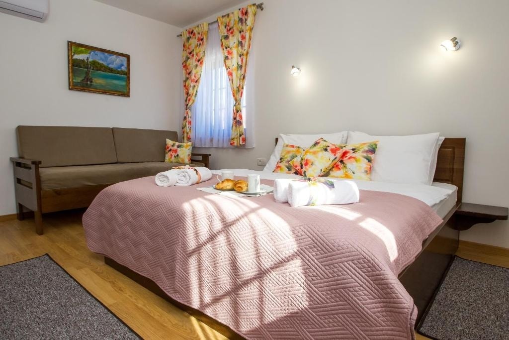 The room features a spacious bed with soft linens, accompanied by a sofa bed. Bright floral curtains frame the window, allowing natural light to illuminate the space. A small table displays pastries and drinks, creating a welcoming and cozy environment.