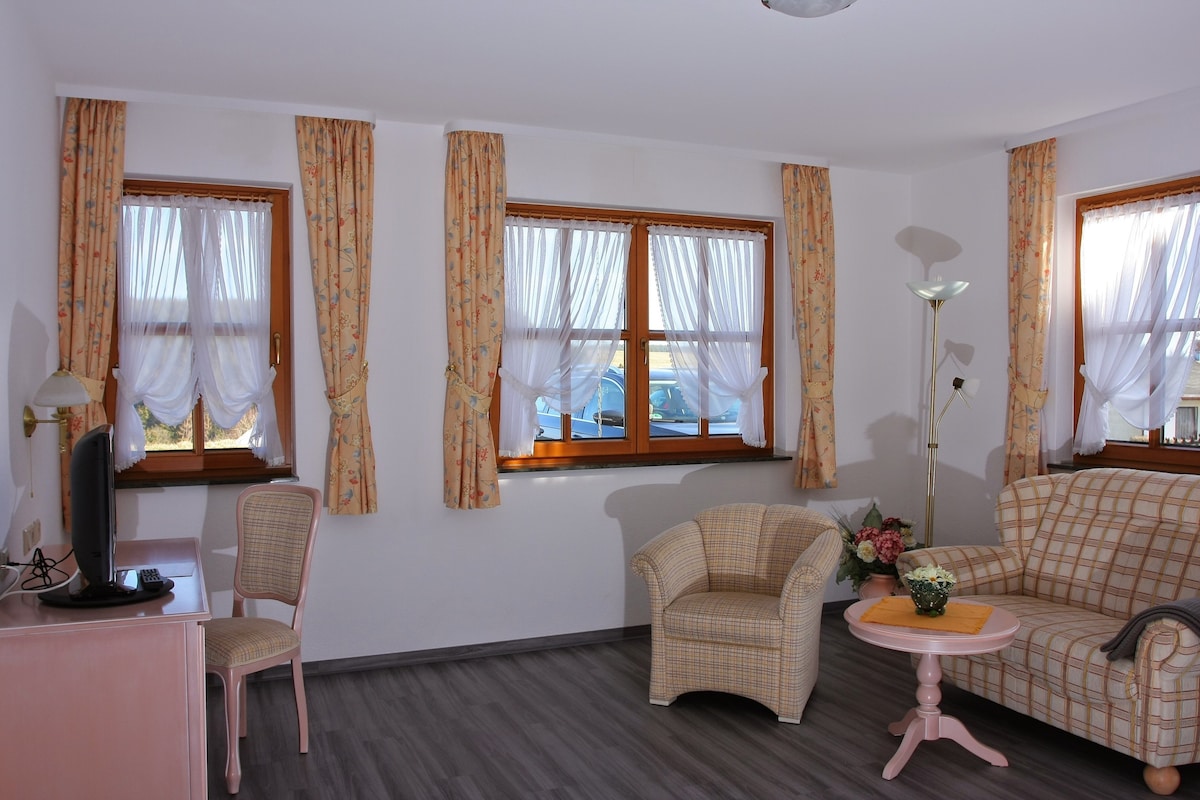 A bright living space features large windows adorned with light curtains, allowing natural light to illuminate the area. A cozy sofa and armchairs are present, accompanied by a small coffee table. A desk and lamp are situated near the corner, offering a functional workspace.