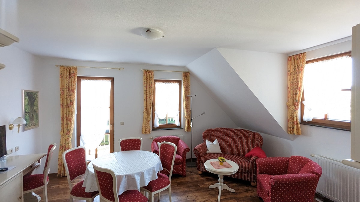 A light-filled living area features a round dining table surrounded by six upholstered chairs. A cozy red couch is arranged alongside another matching seating area. Large windows draped with floral curtains allow natural light to brighten the room, enhancing the warm atmosphere.