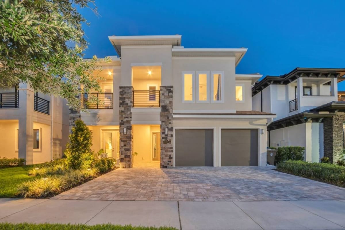 The exterior of a modern villa is displayed, featuring a balanced design with natural stone accents and large windows. A symmetrical driveway leads to two garage doors. The soft glow of warm lighting is visible inside, enhancing the welcoming ambiance as dusk sets in.