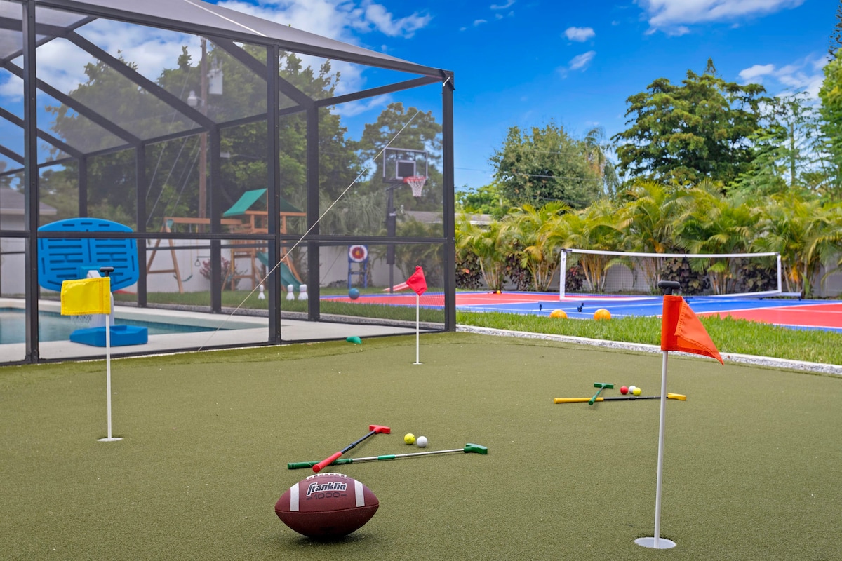 The image features a vibrant outdoor area with a mini-golf course adorned with colorful flags. Nearby, a basketball hoop is visible alongside a swimming pool, and a children's playground is partially visible in the background. Lush greenery surrounds the space.