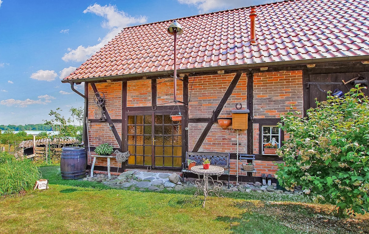 The exterior of a rustic-style barn is showcased, featuring a red tiled roof and a combination of brick and timber elements. A small garden area is visible, with greenery and a decorative barrel. A table and chairs sit invitingly in front of the barn.