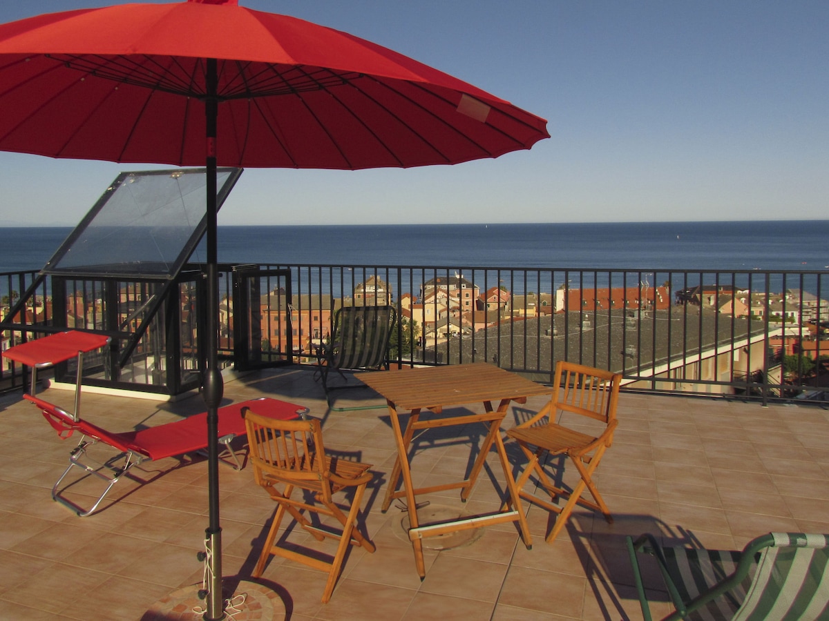 A private roof terrace features wooden and metal furniture, including two lounge chairs and a table. A large red umbrella provides shade, while views of the sea and surrounding landscape are visible in the background, creating a serene outdoor space.