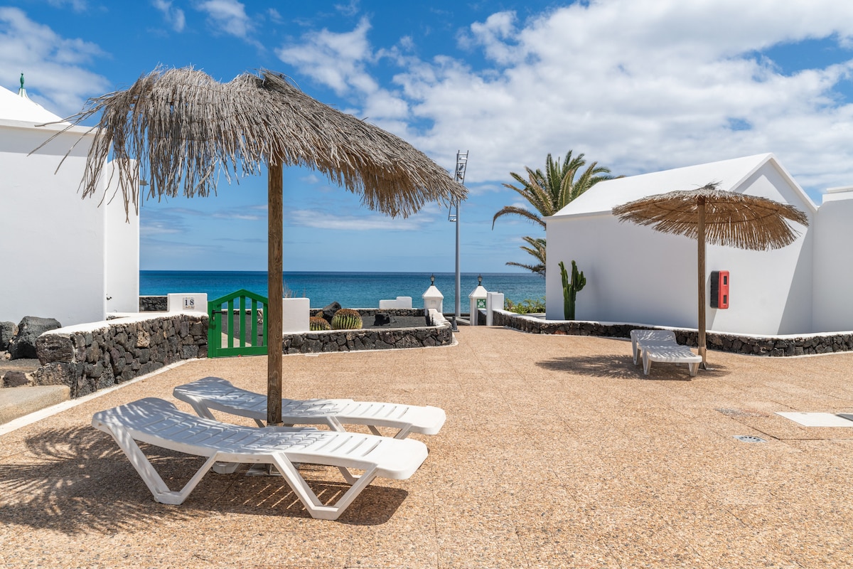 Two lounge chairs are positioned under straw umbrellas on a sunlit patio. The area features a stone wall and a green gate in the background, with the serene blue of the Atlantic Ocean visible beyond. Palm trees and white structures add to the tranquil ambiance.