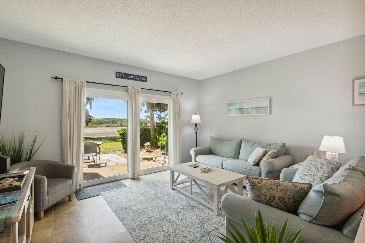 A bright living area features two comfortable sofas and a coffee table, set against a backdrop of natural light streaming in through sliding glass doors. A serene view of the outdoor landscape is visible, with greenery and a walkway just outside.
