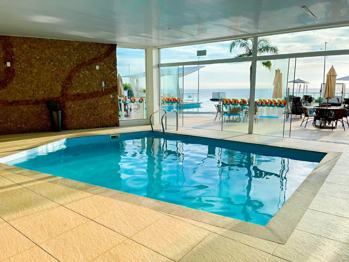 An indoor pool offers a serene setting, with crystal-clear water reflecting light. Large glass walls provide a view of the outdoor area, showcasing additional seating and umbrellas by the infinity pool just beyond.