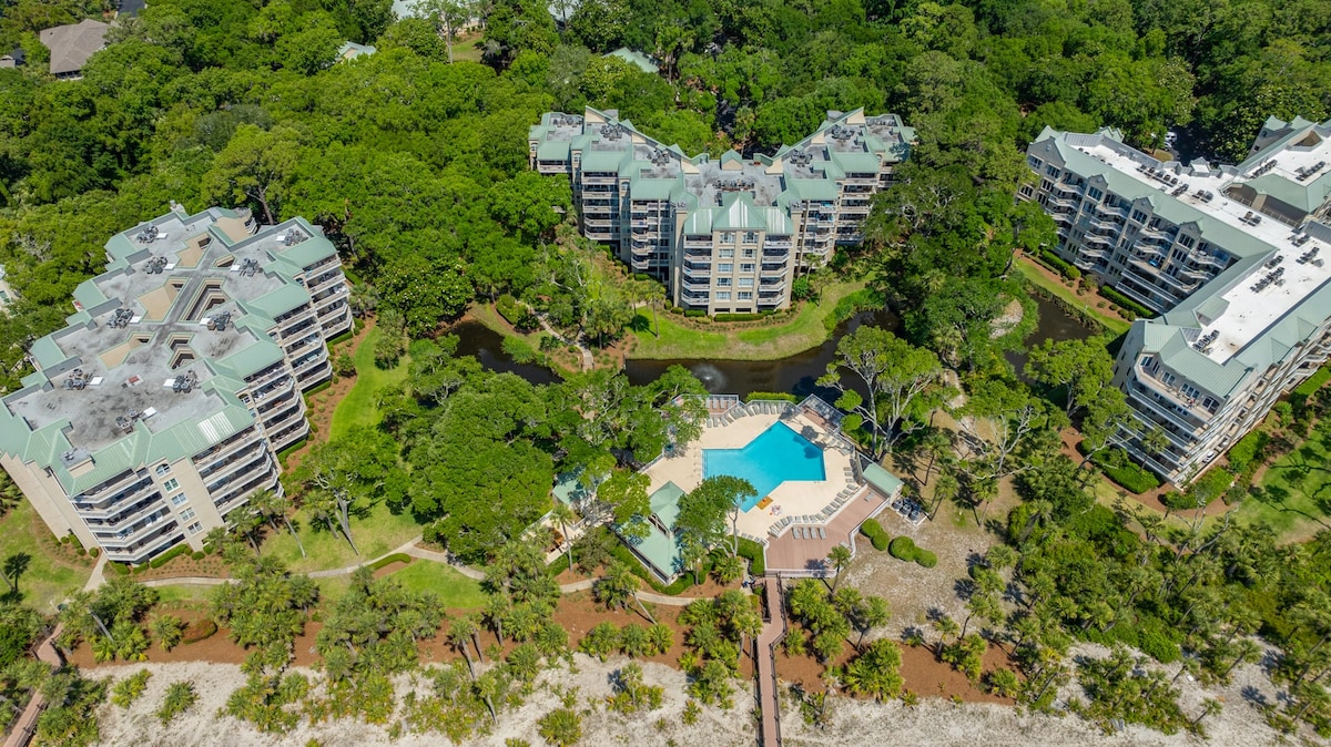 An aerial view captures the layout of the condo complex surrounded by lush greenery. A central pool area is highlighted, flanked by well-maintained landscaping. The buildings feature multiple structures with green roofs, set against a backdrop of trees and small water features, creating a peaceful ambiance.