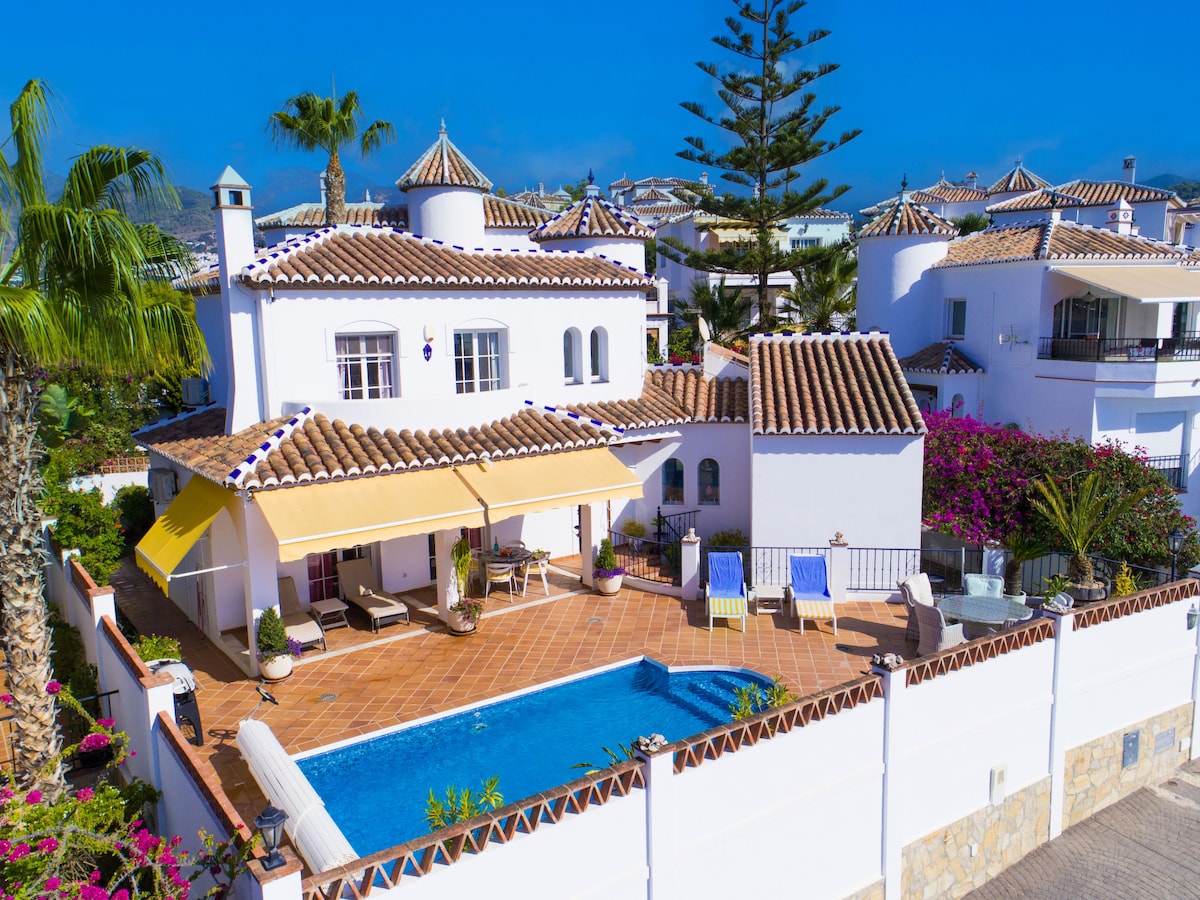 A spacious villa with a private swimming pool is surrounded by a well-maintained garden. The outdoor area features a shaded patio with sun loungers, and vibrant blooming flowers enhance the sunny atmosphere. The architecture showcases traditional Mediterranean elements with a red-tiled roof.