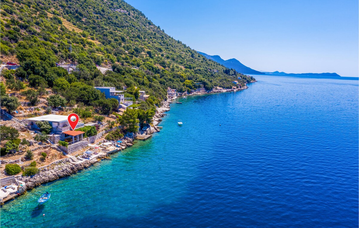 The image captures a semi-detached house positioned along a serene coastline. The crystal-clear water reflects shades of blue, with gentle waves lapping against a concrete plateau. Nearby, boats can be seen floating, while green hills rise in the background, adding to the tranquil scenery.