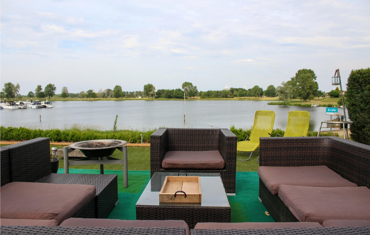 An outdoor seating area is presented with comfortable wicker furniture arranged around a central table. Two lounge chairs are positioned on the left, and a fire pit sits atop a grassy area, with views of the lake visible in the background.