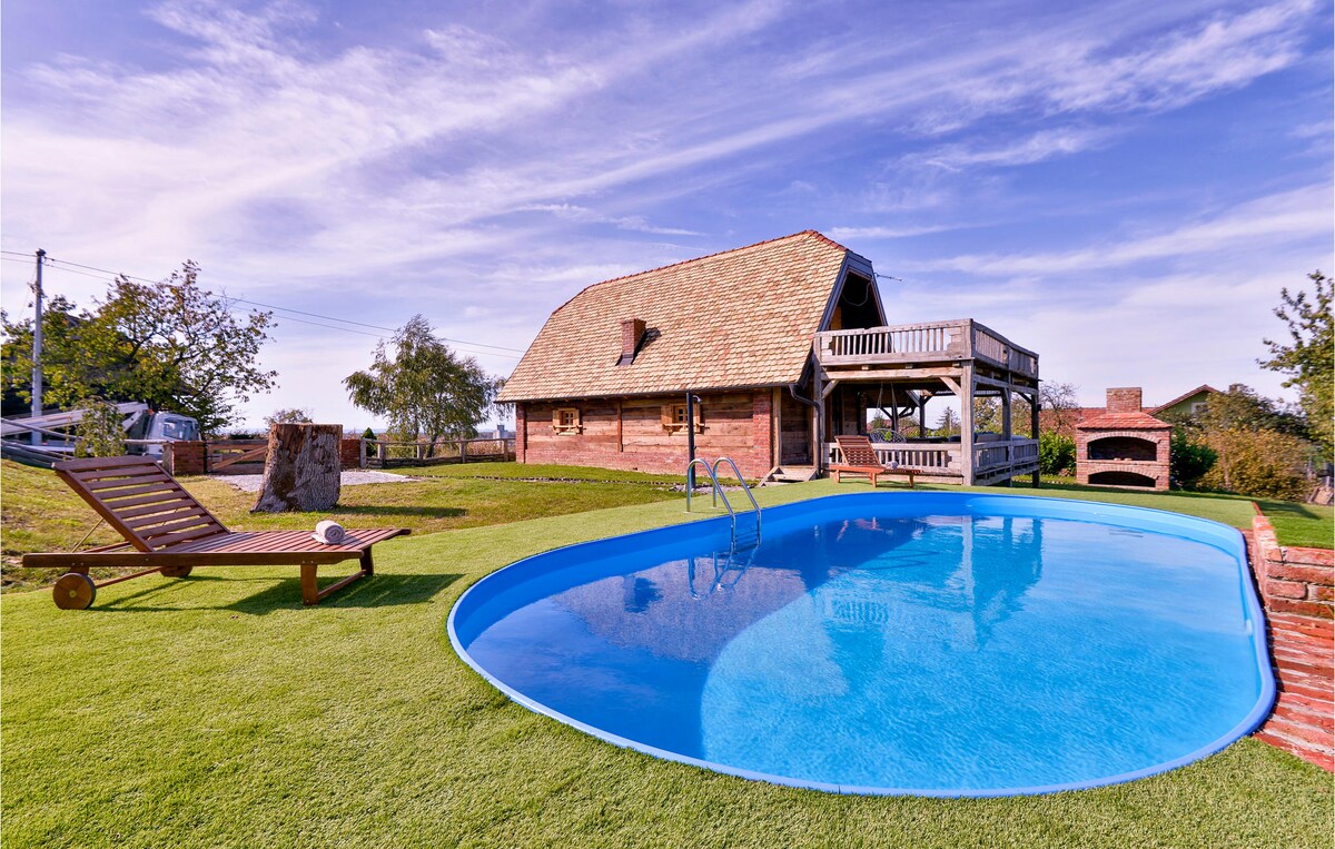 An inviting outdoor space is highlighted by a blue swimming pool, surrounded by lush green grass. A wooden lounge chair is positioned on the lawn, with the charming wooden house and a brick barbecue visible in the background. A bright sky enhances the serene atmosphere.