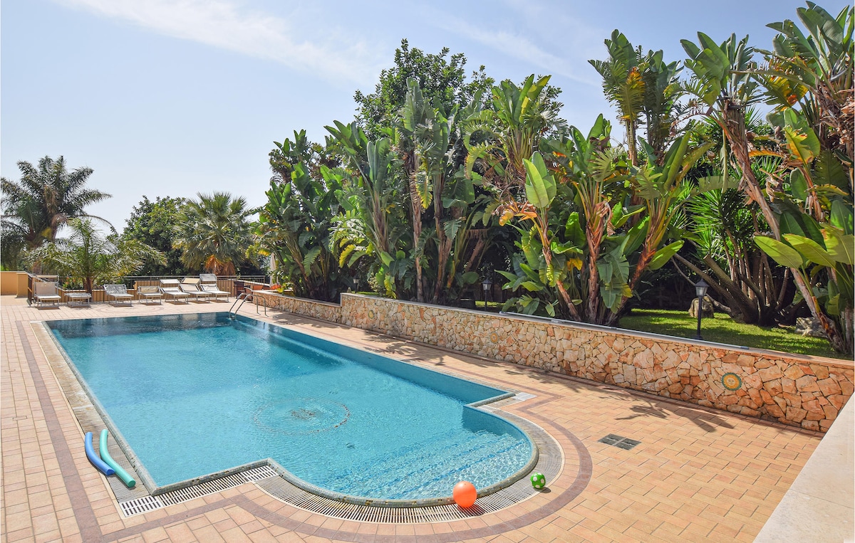 The outdoor pool area is surrounded by lush tropical foliage and palm trees, providing a tranquil environment. The clear blue water is complemented by a spacious deck featuring lounge chairs. A colorful inflatable is visible in the pool, adding a playful touch to the scene.