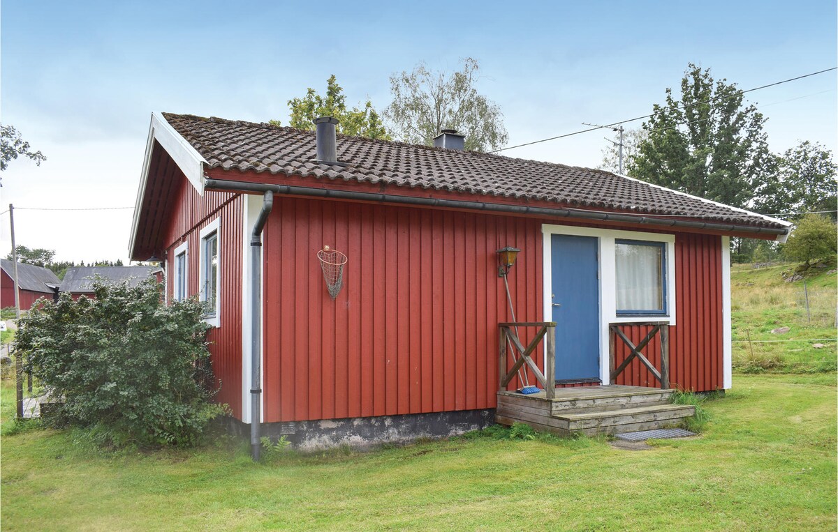 A red cottage with white trim is shown surrounded by green grass and shrubs. The front entrance is accessible via a small set of stairs. Two visible chimneys are located on the roof, and trees provide a natural backdrop.