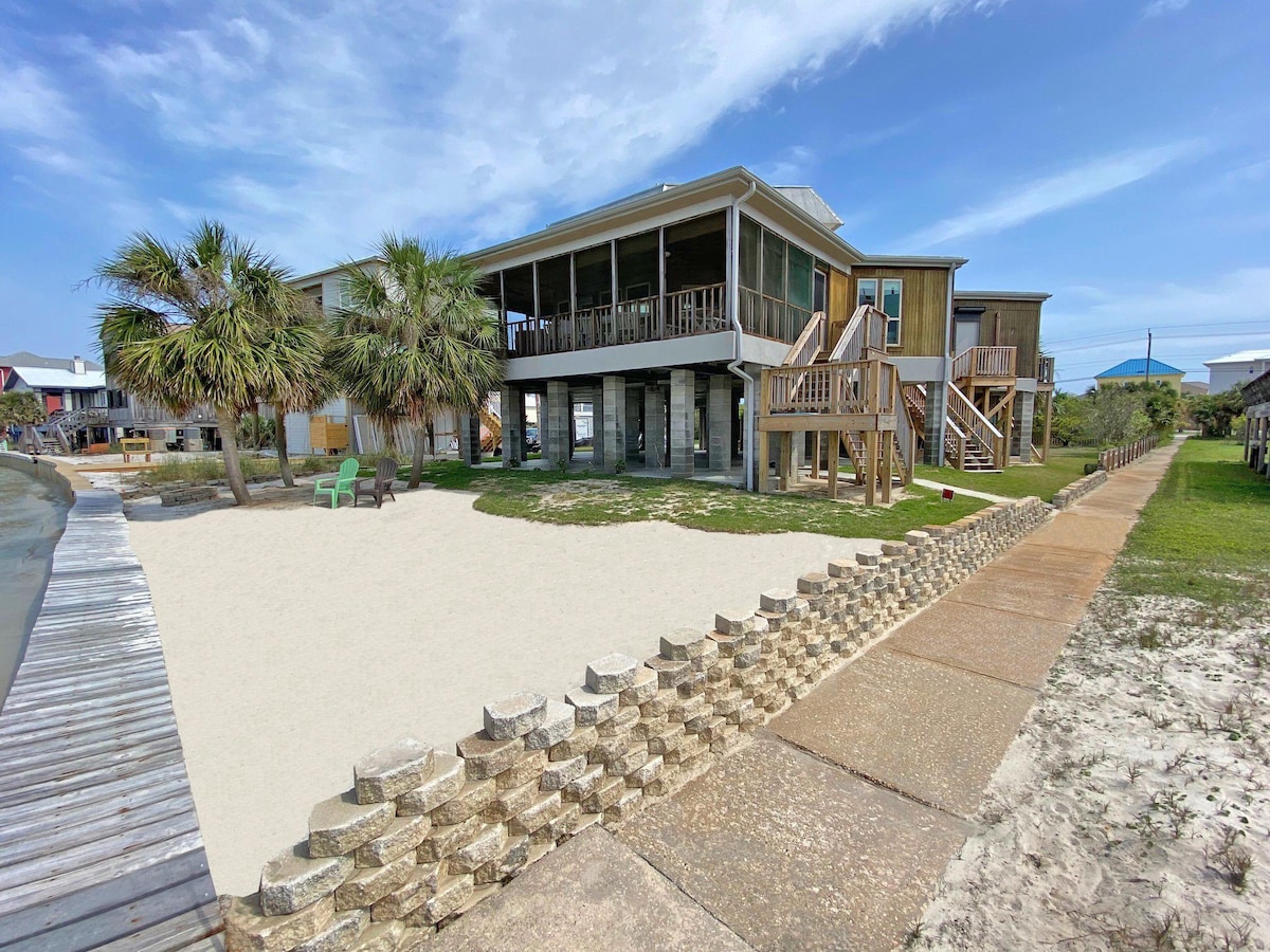 The exterior of the home showcases a spacious sandy area bordered by a low stone wall. Green palm trees frame the property, while steps lead up to the screened-in porch. A wooden pathway runs along the side, offering easy access to the beach and surrounding areas.