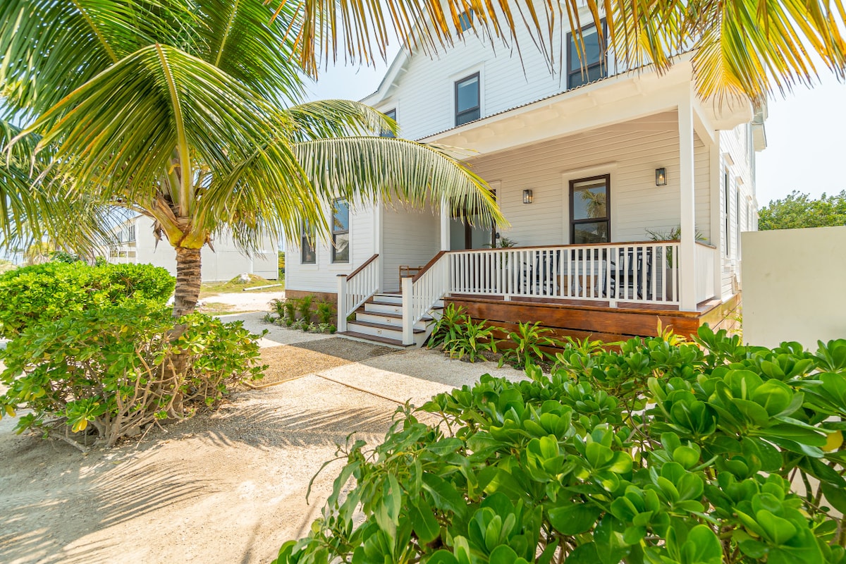 The exterior of the villa is framed by lush green foliage and palm trees. A welcoming front porch with white railings is visible, leading up to a light-colored house with multiple windows, set against a clear sky.