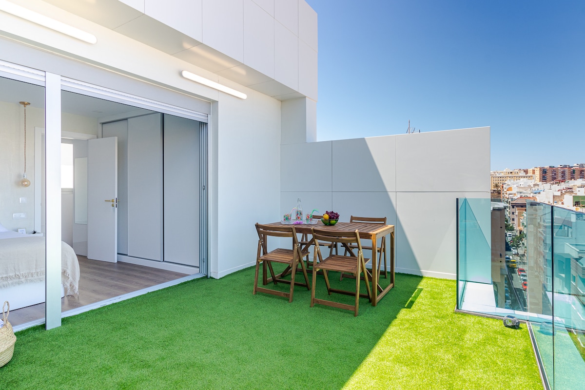 A terrace area features a dining table surrounded by wooden chairs, set against lush green turf. Glass railings provide safety while maintaining views of the urban landscape. Sliding glass doors lead to the interior space, enhancing the connection between indoor and outdoor environments.