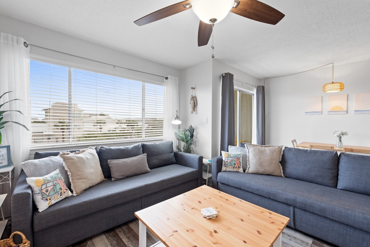 A light-filled living area features two spacious sofas adorned with decorative pillows, arranged around a wooden coffee table. A ceiling fan hangs above, and large windows provide natural light, revealing a view of the outdoors. A dining area is visible in the background.