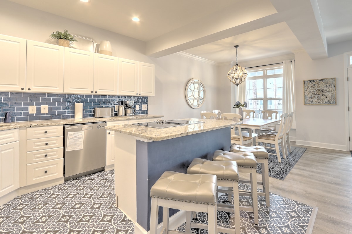 A modern kitchen showcases white cabinetry and stainless steel appliances, complemented by granite countertops and a breakfast bar with four padded stools. A dining area with a round chandelier and a decorative mirror sits adjacent, while intricate tile flooring adds texture and style to the space.