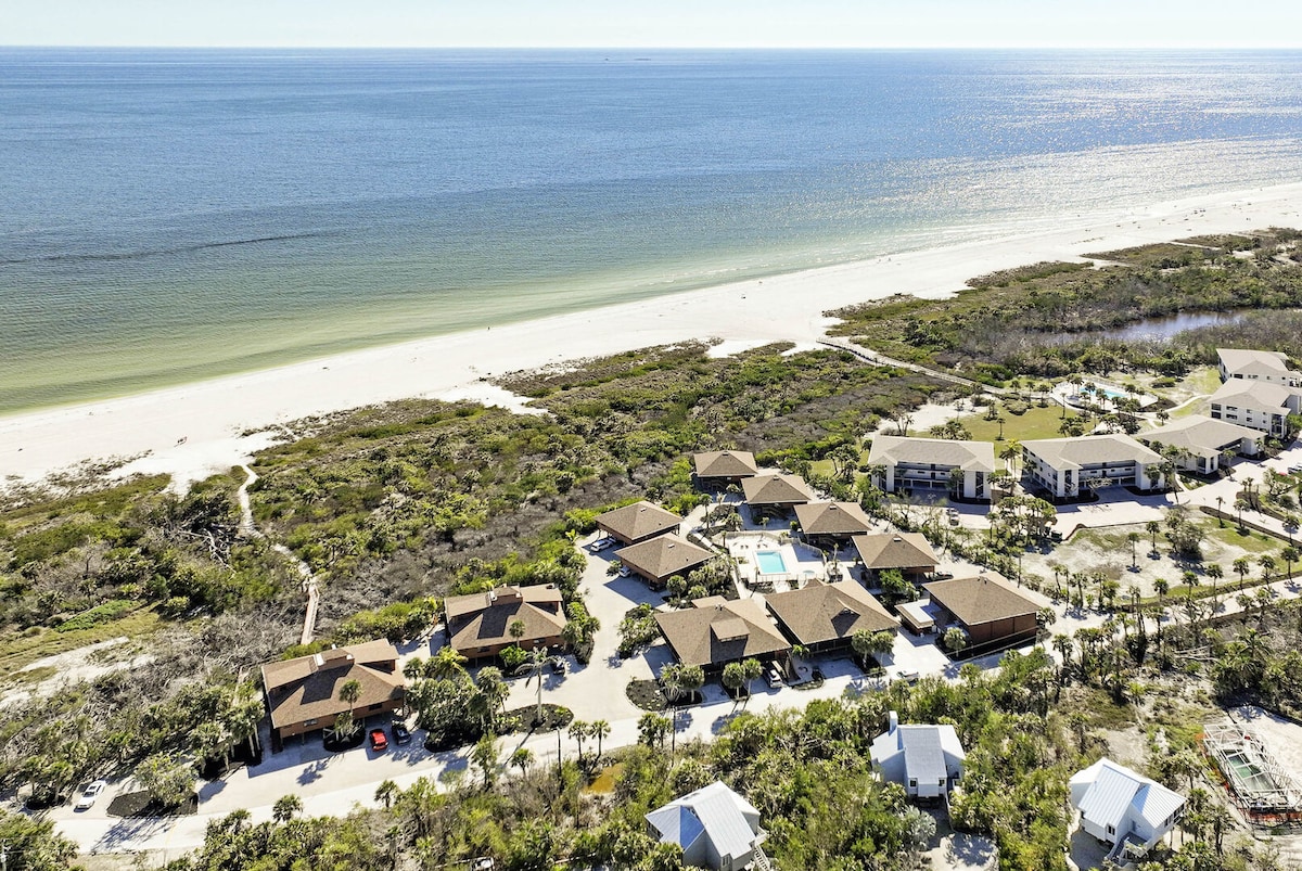 An aerial view captures a cluster of beachside buildings interspersed with lush greenery, situated along a tranquil shoreline. The expansive blue ocean stretches towards the horizon, with gentle waves lapping at the sandy beach. A pool area is visible among the buildings, inviting relaxation.