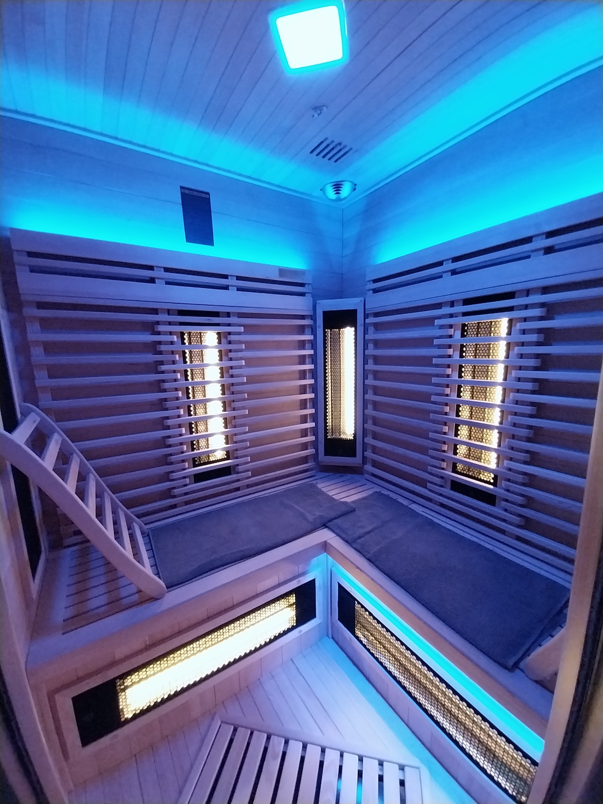The sauna space is designed with a modern aesthetic, featuring smooth wooden panels and subtle blue lighting. Benches are arranged on different levels, providing ample seating. Illuminated panels radiate warmth, creating a calming atmosphere suitable for relaxation.