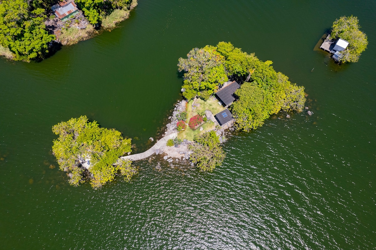 An aerial view displays a tranquil island surrounded by water, featuring lush greenery and vibrant flowers. A narrow path leads to a charming glass house nestled among trees, offering a sense of serenity and privacy within the natural landscape.