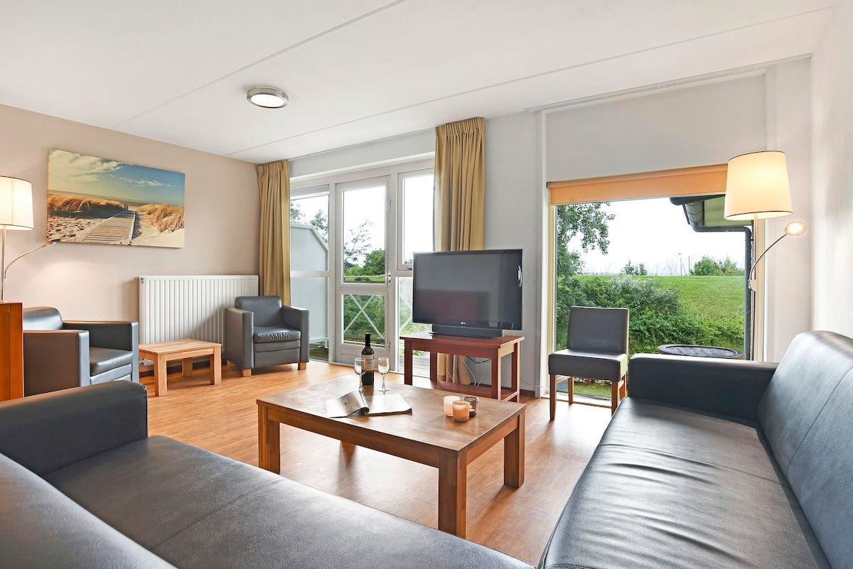 The living room contains a seating area with two dark sofas facing a wooden coffee table. A television sits on a stand opposite the seating. Large windows provide views of the patio outside, while several lamps offer soft lighting.