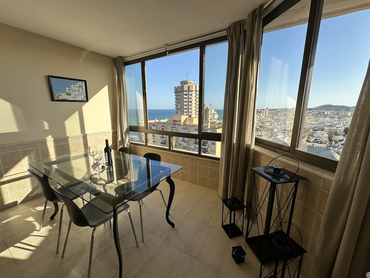 A dining area is presented with a glass table and transparent chairs, set below large windows that reveal views of the sea and city. Natural light floods the space, and a small shelf stands beside the window, enhancing the room's functionality.