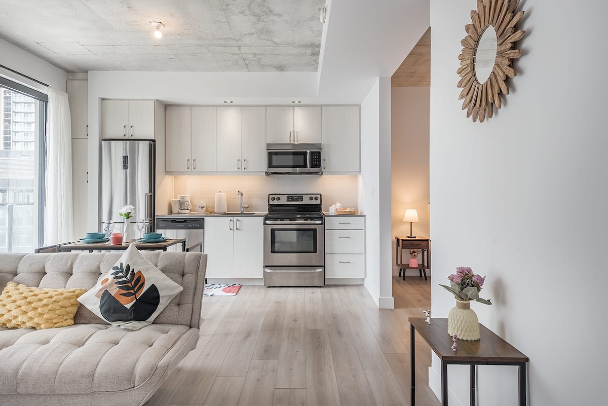 A modern kitchen and living area showcase sleek cabinetry and stainless steel appliances. A comfortable sofa with decorative cushions faces a small dining table, while natural light floods the space through large windows. A mirror on the wall adds a touch of elegance.